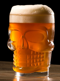Photo of a clear, skull-shaped mug filled with rich, vibrantly orange-hued beer and capped with an overflowing amount of cream colored foam