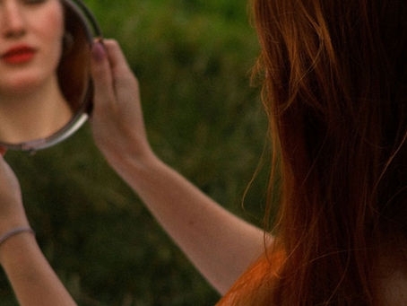 A woman studying her reflection in a mirror representing hormonal skin and hair changes after 40 and the story of Margaux from the Land of Crowns and Chaos by the Happy Juice Chick.