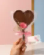 Paleta de chocolate en forma de corazón envuelta en celofán con cinta rosa, flor decorativa y tarjeta del Día de la Madre