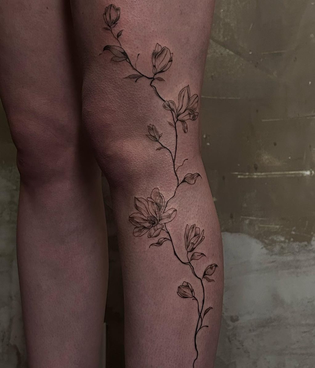 Trailing magnolia branch with open blooms and buds, tattooed across the back of a knee and calf