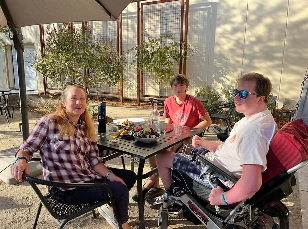 Caregiver and two young men, one using a power wheelchair, seated together at an outdoor restaurant table during a shared meal.