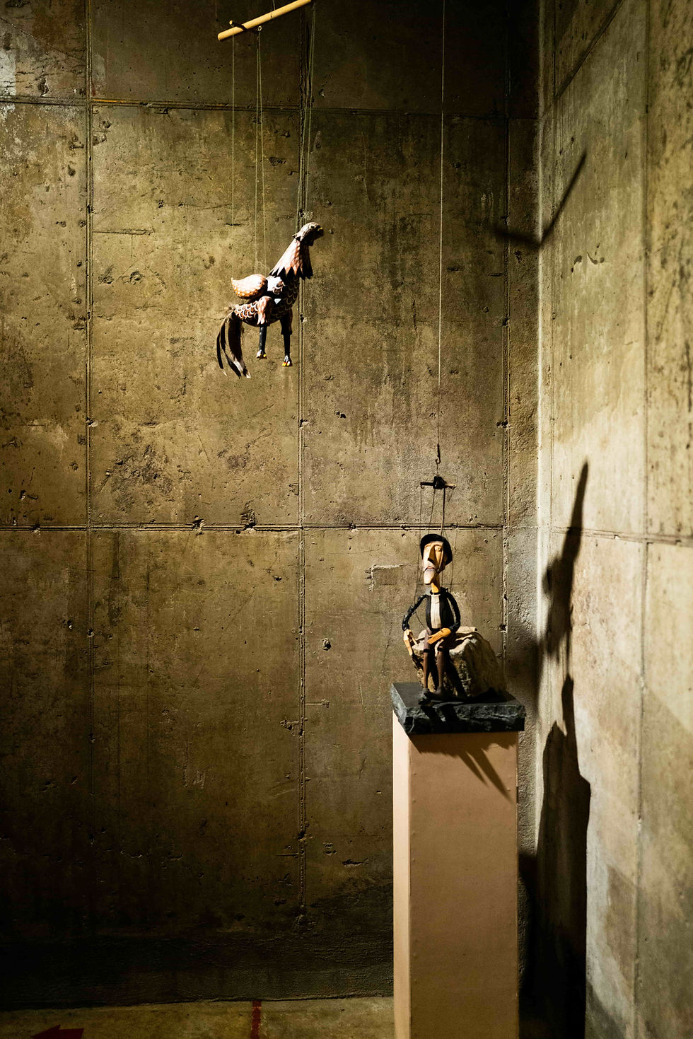 Marionette horse suspended mid-air and a seated marionette on a pedestal in a dim room with concrete walls. Shadows cast on the walls.