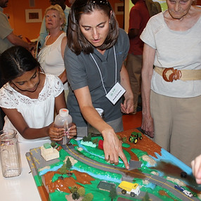 Women pointing at an interactive model while a girl by her side pours some liquid into it.
