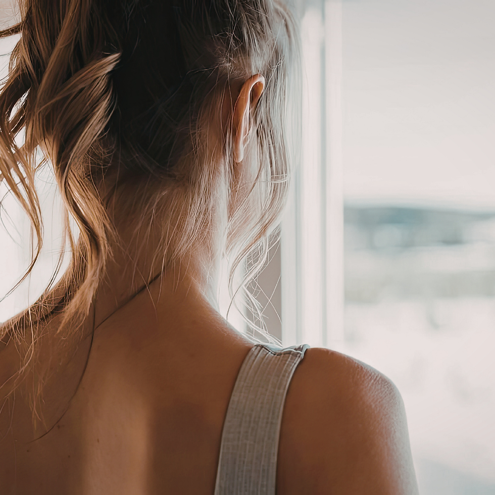 Back view of a woman with a messy bun in a gray tank top, standing by a bright window in soft, calm light.