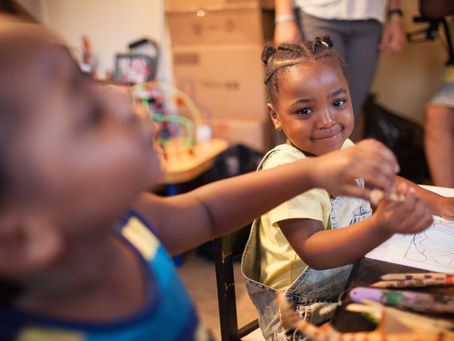 A little Black girl smiles at the camera while drawing a picture.