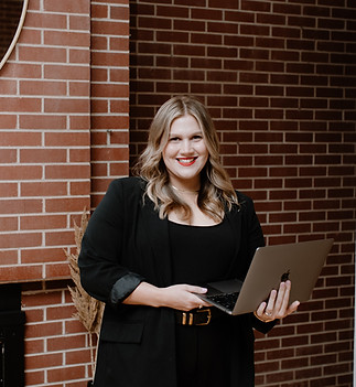 Kenedi, marketing manager at TRU Hair Salon in Colorado Springs, smiling brightly while holding a laptop. She has shoulder-length blonde hair with soft waves and is wearing a black blazer with a black belt. She's standing in the salon's modern interior with exposed brick walls and a round mirror visible in the background. Her professional yet approachable demeanor reflects her role in managing the salon's marketing efforts.
