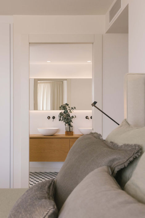 View from bedroom with neutral linen pillows to modern bathroom with double sinks and wooden vanity.