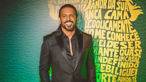 Cantor Thales Lessa posa sorridente com terno preto em evento de lançamento do projeto “O Livro – Do Que Se Sente Pro Que Se Escuta”, em São Paulo, com fundo verde iluminado e painel com títulos de músicas ao fundo.