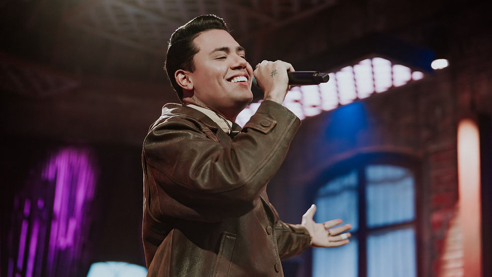 Felipe Araújo cantando em palco com microfone na mão, usando jaqueta de couro, em cenário intimista com iluminação quente e elementos industriais ao fundo.