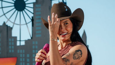 na Castela sorrindo com chapéu e microfone em show na praia.