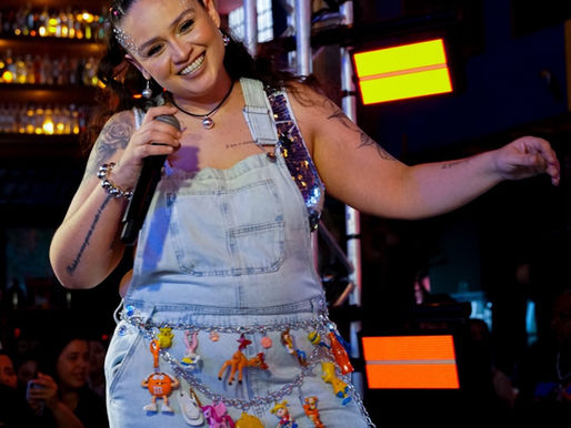 Luiza Martins no palco durante ensaio de Carnaval, sorrindo, segurando o microfone e usando jardineira jeans com adereços coloridos, em clima festivo e descontraído.