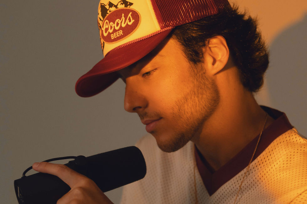 Enzo Ferro segurando um microfone durante sessão de gravação, usando boné vermelho e camisa clara, com iluminação quente e expressão introspectiva.