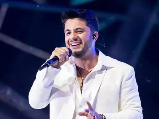 Cristiano Araújo canta ao vivo segurando um microfone, vestindo blazer branco, sorridente, em um palco iluminado por luzes azuis e roxas.