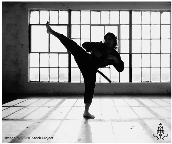 A woman in karate gear standing in a dojo. One foot kicking into the air with the other foot. Her hands are in a defensive position.