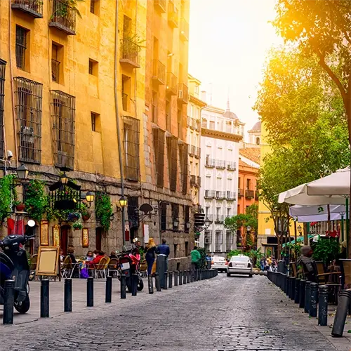 Street view of Cava de San Miguel in Madrid with outdoor cafés, colorful historic buildings, trees, and people enjoying a relaxed atmosphere
