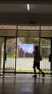 Pessoa caminhando em um corredor interno, vista em silhueta contra a luz externa. Ao fundo, uma fotografia em tecido está instalada em uma área aberta. Acima da passagem, uma placa indica “Saída”.