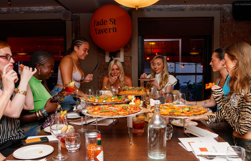 A group of women laughing and enjoying pizza and cocktails around a dining table at Fade St Tavern.