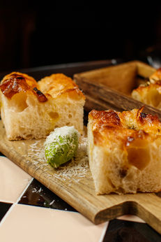 A close-up, eye-level shot of two thick, square slices of golden-brown focaccia bread served on a wooden paddle board. Nestled between the slices is a bright green quenelle of parsley butter, heavily dusted with finely grated parmesan cheese that scatters onto the board, all resting on a black and cream checkered tabletop.