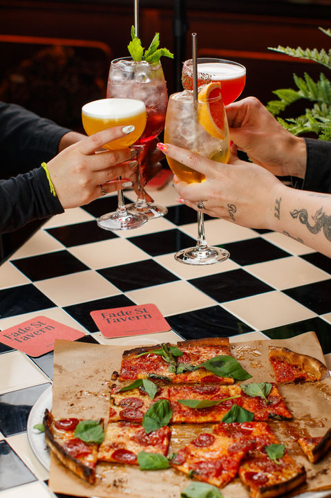 A stylized photograph showing a group of friends bringing their cocktail glasses together for a toast over a table. In the background, a pizza cut into square slices sits on a piece of parchment paper over a checkered tiled tabletop. Several colorful drinks, including a yellow cocktail with a foam top and a tall glass with an orange slice, are held in mid-air by hands, one of which shows a tattoo on the forearm.