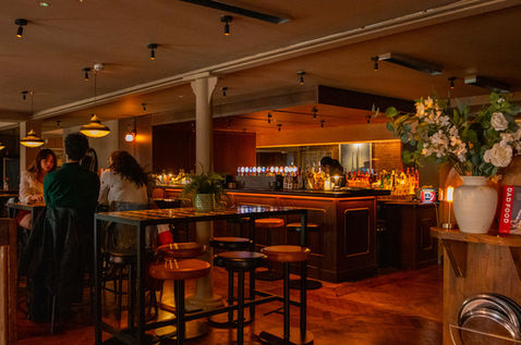 The warmly lit interior of a modern restaurant and bar. In the foreground, high-top dark wooden tables are surrounded by matching wooden barstools and leather-backed chairs. In the mid-ground, a long bar counter with beer taps is visible, with bartenders working behind it. Pendant lights with a warm amber glow hang from the ceiling, illuminating the cozy dining space.