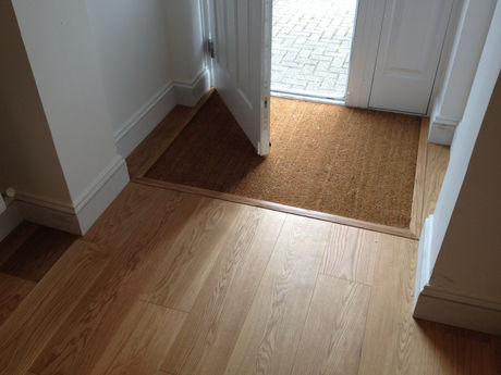 Empty hallway with light wood floor and open white door, sunlight on a brown doormat leading to a brick patio.