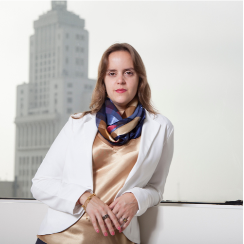 Mulher de blazer branco e lenço colorido posa séria diante de prédio alto em fundo desfocado.