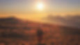Person in a coat walks on a grassy hill during sunrise, casting long shadows. Warm orange hues fill the sky, with distant mountains. EMDR Therapy at Cadence Psychology Studio in Fishers, Indiana.