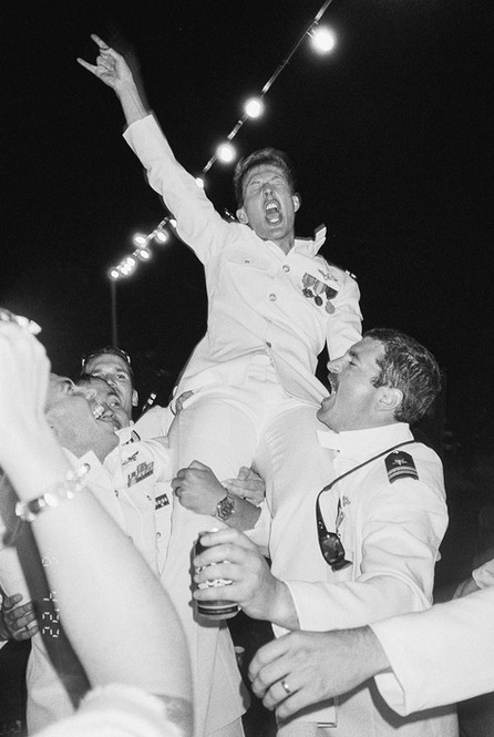 Air Force pilots, wearing dress whites, lift a groom in celebration at a string light lit wedding reception. Image is grainy and shot on black and white 35mm film in San Diego, CA.