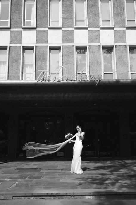 Black and white film portrait of a bride standing outside The Dewberry wedding venue in Charleston, SC. Her veil is caught in a strong gust of wind, blown across her body as she laughs in surprise. 