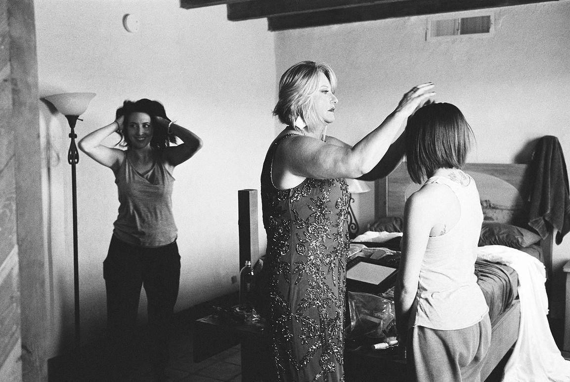 Grainy, black and white, documentary style wedding photograph on 35mm film of a bride and family members doing their hair inside a stucco retreat home in West Texas
