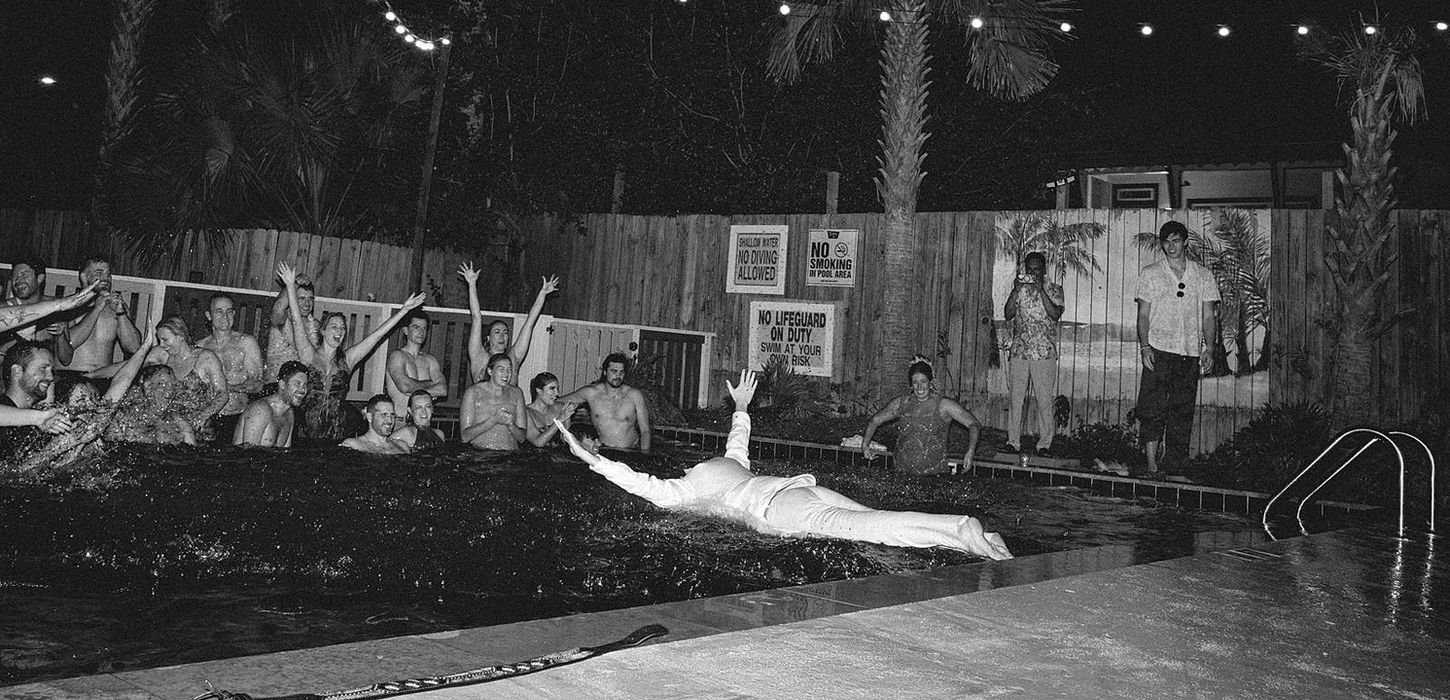A guest just breaks water as he dives into a pool with other wedding guests in this black and white, documentary wedding photograph on Folly Beach.