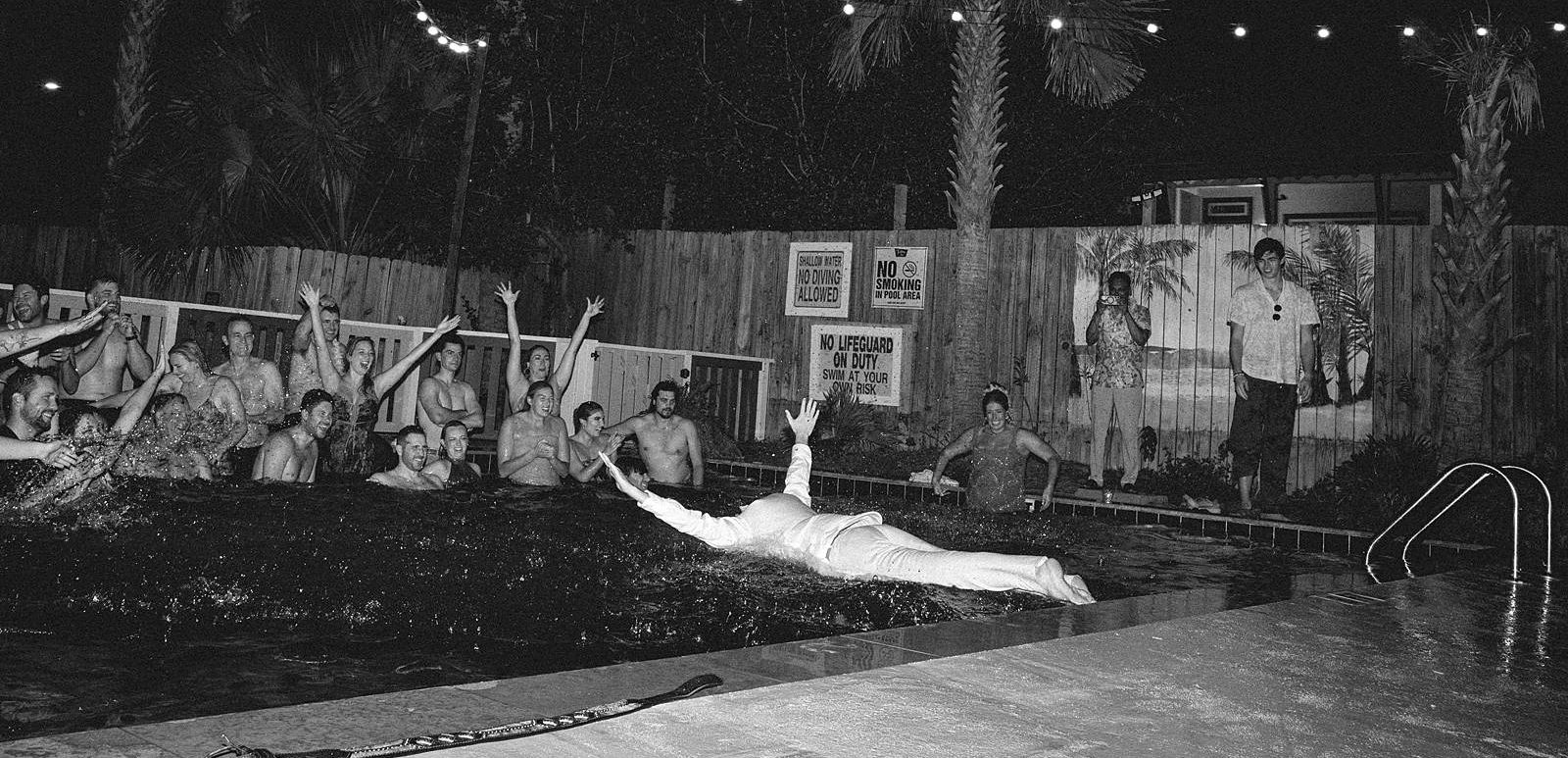 A guest just breaks water as he dives into a pool with other wedding guests in this black and white, documentary wedding photograph on Folly Beach.