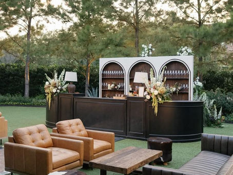 A relaxed and stylish Father’s Day outdoor gathering setup featuring a lounge-style seating arrangement with leather chairs, wooden tables, and soft, earthy-toned textiles. Grilled food is served buffet-style with hearty meats and seasonal sides, while simple greenery, bourbon bottles, and thoughtful personal touches like cigars and handwritten cards create an atmosphere of comfort and appreciation. The setting emphasizes connection, intention, and a laid-back, masculine design that celebrates fathers across generations.
