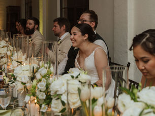 Bride seated at an intimate wedding reception table with guests, candlelight, white florals, and warm neutral décor.