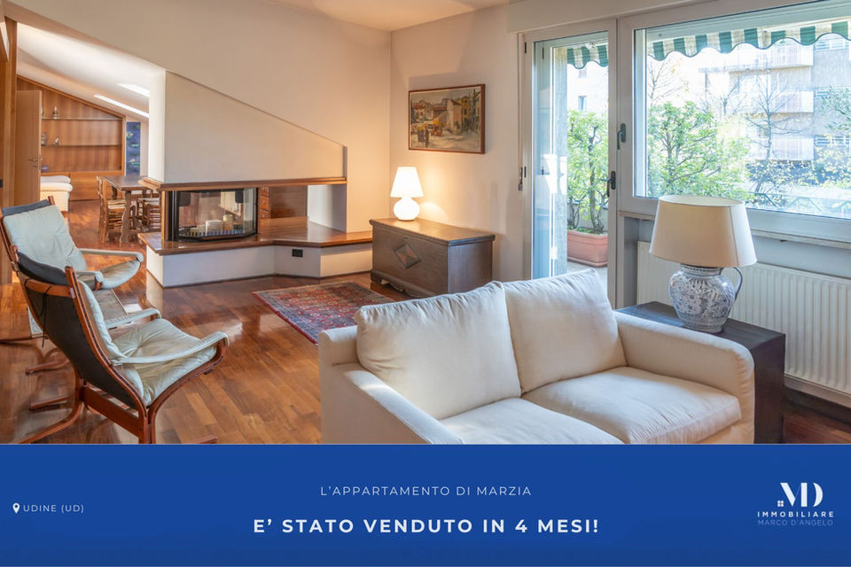 Un'immagine pubblicitaria di Immobiliare Marco D'Angelo che mostra un moderno e accogliente salotto di un appartamento a Udine. La foto cattura un moderno camino, due poltrone di design, un divano bianco e una grande finestra che si apre su un balcone con piante. In basso, una fascia blu contiene il testo: "L'APPARTAMENTO DI MARZIA E' STATO VENDUTO IN 4 MESI!", con "UDINE (UD)" a sinistra e il logo "MD IMMOBILIARE MARCO D'ANGELO" a destra.
