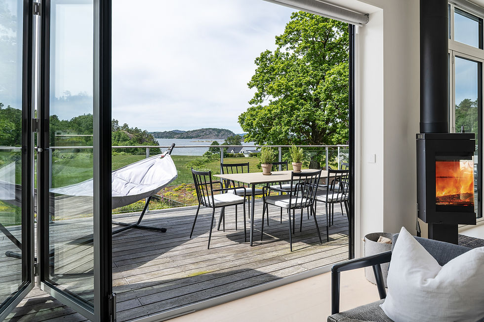 Uteplats med matbord, stolar och hängstol mot sjövy; öppen brasa inomhus och grönskande träd.