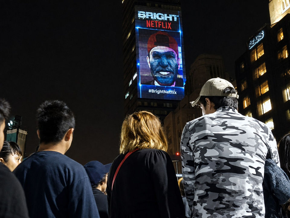 2 jóvenes y su madre frente a torre latino ven una video proyección sobre la fachada en la que se ve el rostro de un chico transformado en arco mediante un filtro digital