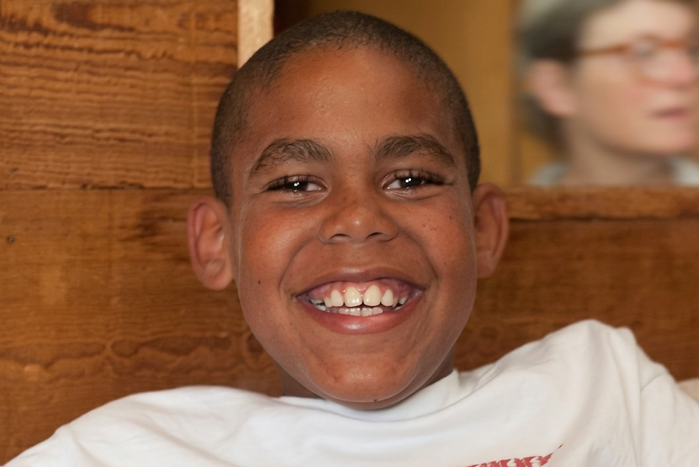 Studio photography of a young boy smiling broadly at the camera. The setting features warm wooden paneling in the background, creating a friendly and casual atmosphere.