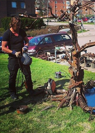 A candid shot of sculptor Olav Lunde on-site, providing a sense of scale for the mechanical tree. He is positioned near the base of the sculpture, surrounded by tools and raw metal materials.