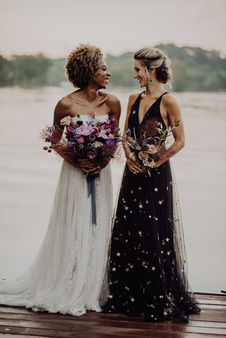 Two brides smiling by a lake in bridal gowns during a wedding at Cherokee Dock. Photographed by Wilde Company.