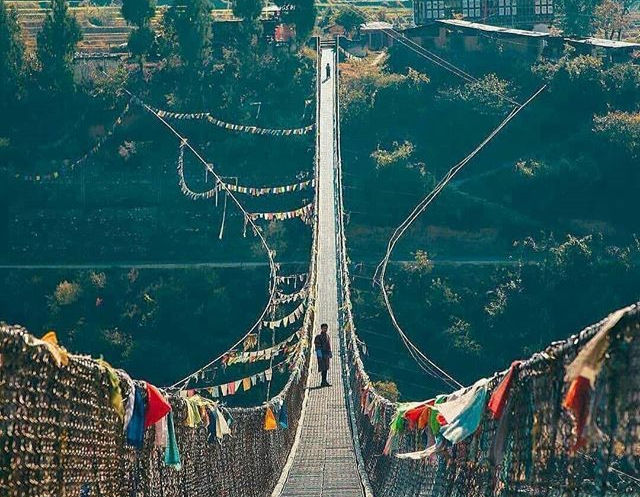 Lone person walks a narrow suspension bridge draped with colorful prayer flags over a deep green valley.