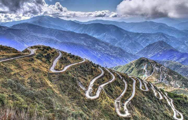 Winding mountain switchback road on steep green ridges under dramatic clouds, with layered blue peaks in the background.