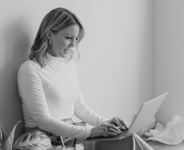 Désirée Tobler sitzt auf einem Kissen am Boden und arbeitet an einem Laptop. Neben ihr steht eine Zimmerpflanze, im Hintergrund sind eine helle Wand, ein Fenster mit Vorhang zu sehen.