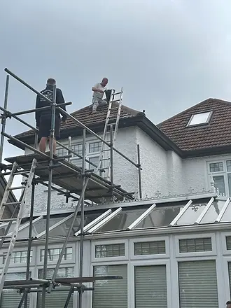 Two workers on scaffolding perform maintenance on a roof, one standing on a ladder and the other on the roof of a conservatory