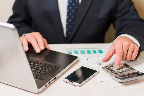 A suited person uses a calculator and laptop on a desk cluttered with papers and a smartphone
