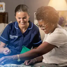 A caregiver in a blue uniform and an elderly woman are smiling while playing Scrabble