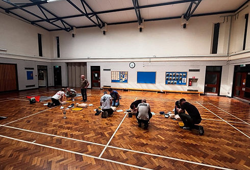 People practicing CPR on mannequins in a large gymnasium with a wooden floor