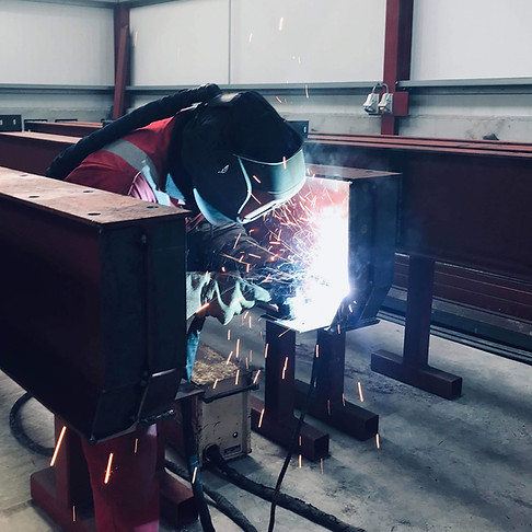 A welder wearing protective gear is focused on welding a large metal beam in an industrial setting