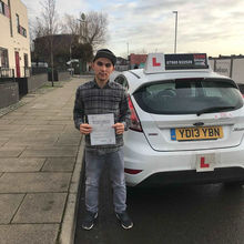 Young man standing on a sidewalk next to a white car with L plates, holding a certificate