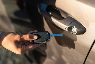 A hand holds a remote car key near a brown vehicle's door handle, sunlight casting a shadow